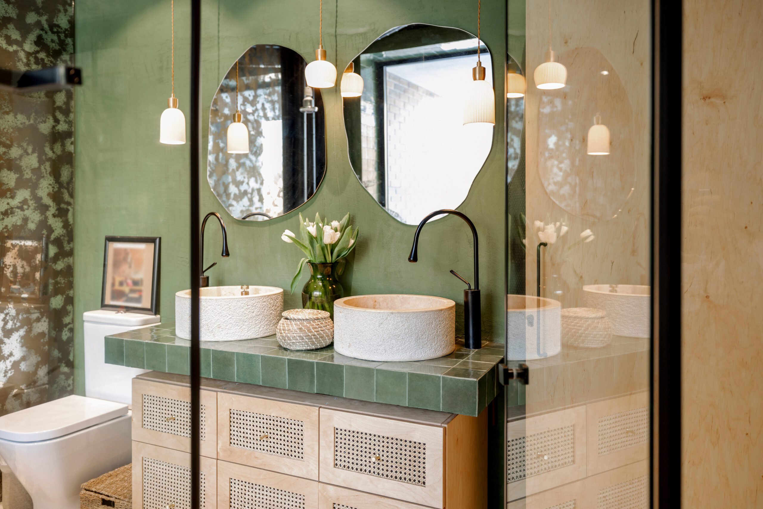 Bathroom interior in natural boho style Bathroom interior with stone wash basins, modern faucets, irregularly shaped mirrors and wooden rattan commode in green and beige tones in natural boho style